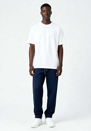 Giovane in piedi che indossa una T-shirt bianca semplice, pantaloni jogging blu navy e scarpe da ginnastica bianche su uno sfondo chiaro.