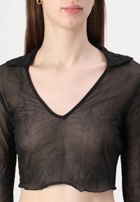 Svart, gjennomsiktig, langermet crop-top med V-hals og volangkant. Stoffet ser rynkete ut, med en kontrasterende svart kant rundt utringningen.