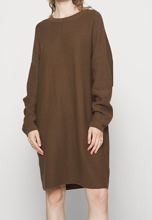 Kobieta ubrana w luźną, długą, brązową, prążkowaną, dzianinową sukienkę swetrową stoi na tle jednolitego jasnego tła.