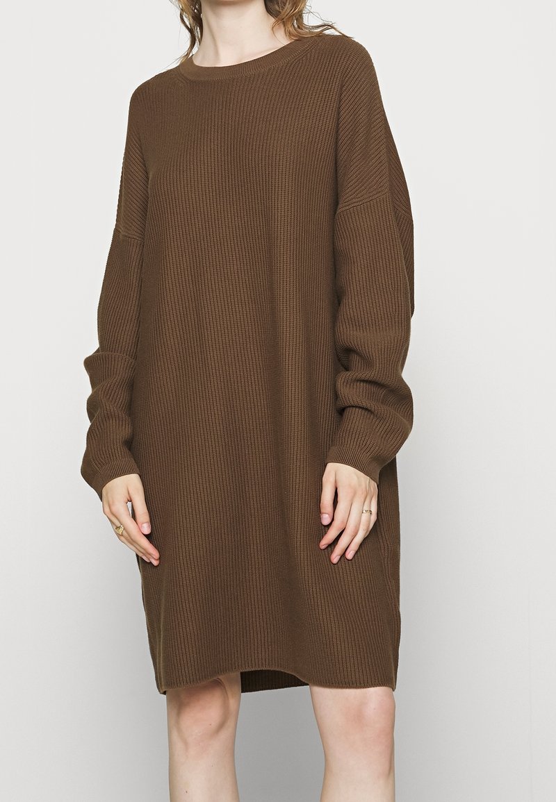 Femme portant une robe-pull en tricot côtelé marron, ample et à manches longues, debout devant un fond clair uni.