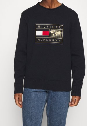 Czarny sweter z prostokątną naszywką z napisem „HILFIGER” nad mapą świata oraz czerwonymi i białymi blokami kolorystycznymi, noszony z niebieskimi dżinsami.