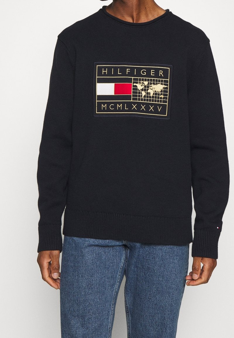 Pull noir avec un patch rectangulaire affichant le texte "HILFIGER" au-dessus d'une carte du monde et des blocs de couleur rouge et blanche, porté avec un jean bleu.