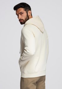 Felpa crema con cappuccio regolabile, tasca a marsupio, polsini a costine e orlo a costine. Indossata con pantaloni marrone chiaro, vista di spalle.