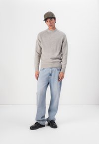 Helles graues Strickpullover mit rundem Ausschnitt, kombiniert mit ausgebleichten blauen weit geschnittenen Jeans. Eine braune karierte Mütze und schwarze Schuhe runden das Outfit ab.
