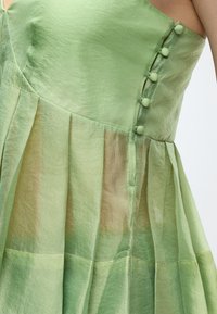 Πράσινο σατέν φορέμα με κουμπιά καλυμμένα με ύφασμα κατά μήκος της πλευράς και λεπτομέρεια πλισέ φούστας στον κορμό ενός ατόμου.