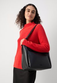 Schwarze Ledertasche mit einem lässigen Design, ausgestattet mit einer großen Öffnung und einem langen Schulterriemen, gehalten von einer Person in einem roten, gerippten Pullover.