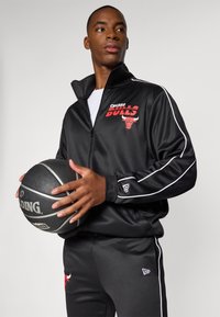 Black tracksuit with red "Chicago Bulls" logo; zip-up jacket with white accents, matching pants, and a black basketball held in hands.