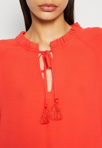 Gros plan sur une femme portant un chemisier rouge avec un col à volants et un lien à franges au niveau du cou.