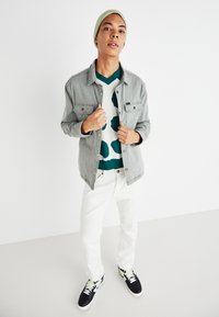 Chaqueta de mezclilla gris sobre un suéter verde con estampado, jeans blancos y zapatillas negras con detalles verdes; modelo con un gorro claro.