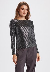 Top lungo nero a maniche lunghe ricoperto di paillettes argentate verticali, con scollatura a barca e vestibilità comoda, abbinato a pantaloni larghi scuri.