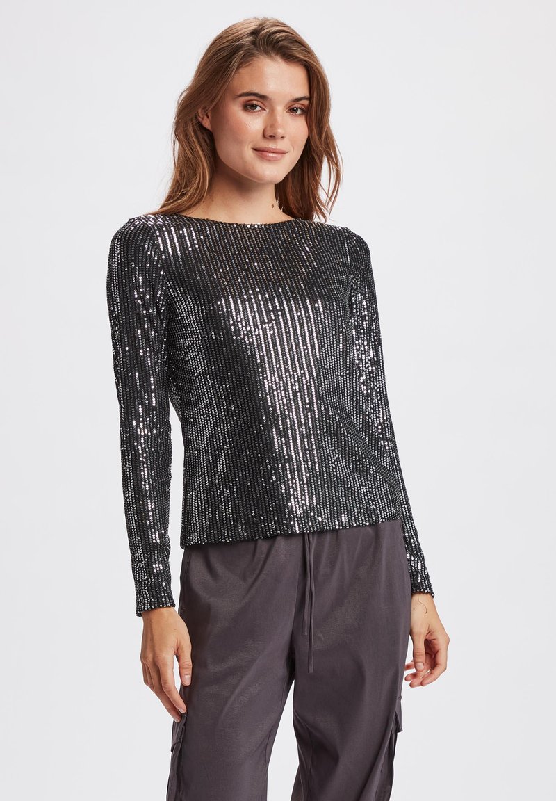 Top lungo nero a maniche lunghe ricoperto di paillettes argentate verticali, con scollatura a barca e vestibilità comoda, abbinato a pantaloni larghi scuri.