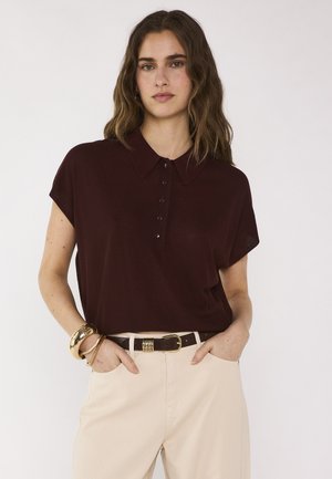 Femme aux cheveux bruns ondulés portant un polo à manches courtes marron foncé, un pantalon beige, une ceinture noire et des bracelets dorés, debout les mains dans les poches.