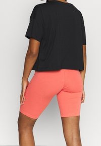 Osoba v korálově oranžových přiléhavých šortkách a volném černém krátkém rukávu, stojící s jednou rukou opřenou o stehno.