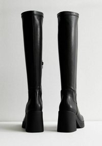 Botas de cuero negras hasta la rodilla con una textura suave, tacón grueso y cierre con cremallera trasera. Presentan un diseño minimalista y punta redondeada.