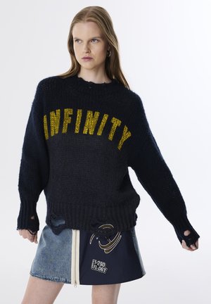 Pull en maille marine avec l'inscription "INFINITY" en lettres jaunes, présentant une coupe ample, des poignets côtelés et des détails usés. Associé à une jupe en jean bleu.