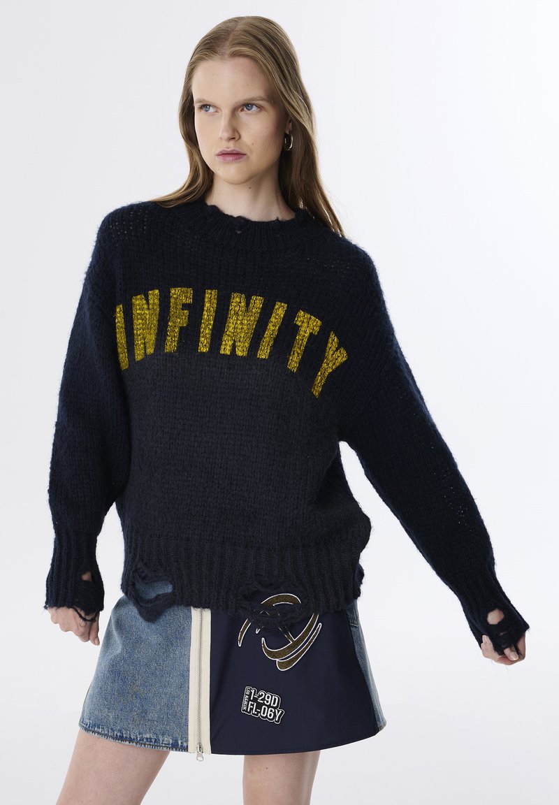Pull en maille marine avec l'inscription "INFINITY" en lettres jaunes, présentant une coupe ample, des poignets côtelés et des détails usés. Associé à une jupe en jean bleu.