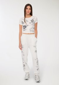 Biała krótka koszulka z czarnymi graficznymi nadrukami, zestawiona z białymi dresami z różnymi rysunkami. Obie części są w luźnym i swobodnym kroju.