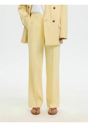 Nő halvány sárga, testhez szabott, széles szárú nadrágot visel, hozzáillő dupla gombolású blézert, fehér felsőt, és barna, nyitott orrú szandált.