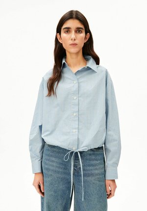 Vrouw met lang donker haar draagt een lichtblauw geruit overhemd met knopen en een gestrikte taille, en blauwe spijkerbroek tegen een effen witte achtergrond.