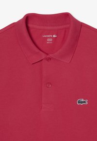 Punainen polo-paita, joka on valmistettu 97 % puuvillasta ja 3 % elastaanista. Ominaisuuksiin kuuluu klassinen kaulus, kaksi nappia ja musta Lacoste-logo vasemmassa rinnassa. Normaalileikkoinen.