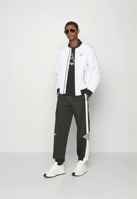 Valge lukuga jope musta graafilise T-särgi peal, koos hallide pükstega, millel on valged ja hallid küljeribad, ning valgete tossudega.