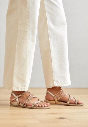 Płaskie sandały na paski w metalicznym złocie o gładkiej fakturze, z regulowanym paskiem wokół kostki, zestawione z jasnymi spodniami o szerokich nogawkach.