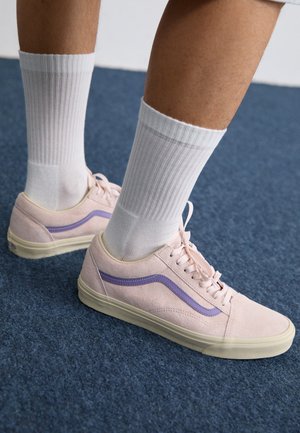 Ben iført hvide ribstrikkede crew-sokker og lyserøde sneakers med lilla sidelister, stående på en blå tæppe.