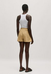 Beige Shorts mit einem strukturierten Muster, elastischem Bund und lockerer Passform. Kombiniert mit einem weißen Tanktop und schwarzen Rutschsandalen.