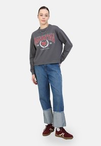 Sudadera gris con gráfico rojo "MANHATTAN", combinada con jeans azules holgados que tienen un dobladillo de denim. Modelo con zapatillas burdeos.