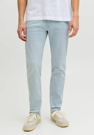 Man draagt lichtblauwe slim-fit jeans, witte sneakers en een wit overhemd, staande tegen een effen lichte achtergrond.