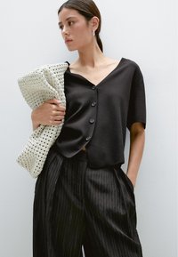 Zwart topje met korte mouwen, een v-hals en vier knopen, gecombineerd met zwarte wijduitlopende broek met krijtstrepen. Houdt een witte geweven clutch met textuur vast.