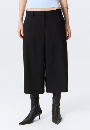 Kobieta w czarnych, szerokich spodniach o długości do połowy łydki oraz czarnych botkach na obcasie z ostrym noskiem, stojąca na jasnym, jednolitym tle.