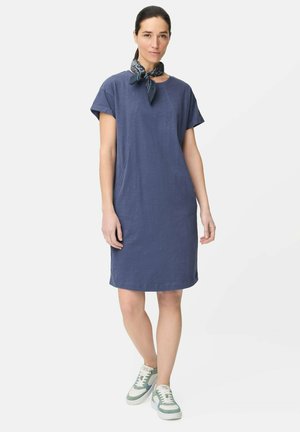 Kvinde iført en knælængde blå hverdagskjole, mønstret halsedisse og hvide og grønne sneakers, stående mod en ensfarvet baggrund.