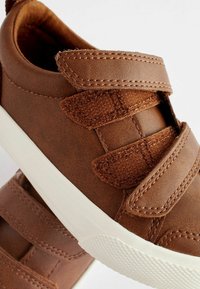 Chaussures en faux cuir marron avec trois sangles Velcro réglables, semelles en caoutchouc blanc et accents en tissu texturé près des sangles.