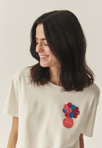 Krémové bavlnené tričko s kvetinovým háčkovaným vzorom v červenej, ružovej, modrej a zelenej farbe, s okrúhlou červenou nášivkou s nápisom.