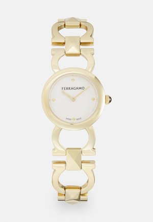 Reloj dorado con una esfera redonda, esfera texturizada blanca y pulsera de eslabones con acentos geométricos. Nombre de la marca "FERRAGAMO" en exhibición.