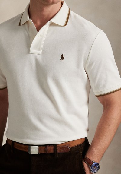 Bílé polo tričko s hnědým límečkem a akcentem. Malé hnědé logo na hrudi. Nošeno s hnědým koženým pásem a modrými hodinkami. Hladká textura.