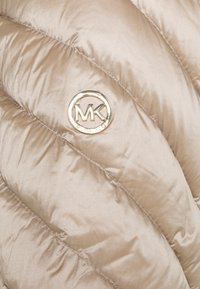 Beige quiltad puffer-väst med en glänsande textur och diagonala sömmar, accentuerad av en guldtonad cirkulär MK-logotyp.