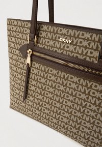 Καφέ τσάντα DKNY tote με μοτίβο λογότυπου, φερμουάρ μπροστινής τσέπης και αντίθετες υφές σε χερούλια και σώμα. Χρυσές λεπτομέρειες.