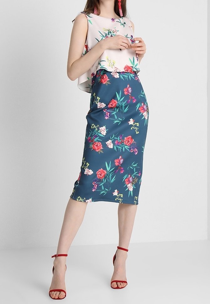 Femme portant un top sans manches à motif floral et une jupe fleurie assortie longueur genou avec des sandales rouges à talons hauts, debout devant un fond uni.