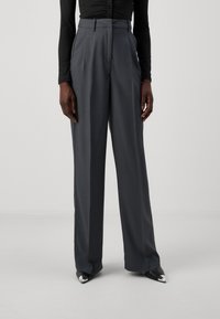Pantalons larges gris en tissu lisse avec taille haute, présentant des plis avant et une coupe ajustée. Assortis à des chaussures noires à bouts pointus.