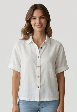 Glunderende vrouw met schouderlang golvend bruin haar, draagt een wit overhemd met korte mouwen en lichtblauwe spijkerbroek, staand tegen een effen achtergrond.