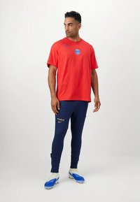 Sarkana Nike T-krekls ar zilu logo, kombinēts ar tumši ziliem sporta biksēm ar zeltainu "PARIS" uzrakstu un zilām futbola kedām.