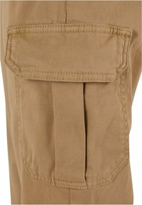 Gros plan sur une poche cargo en tissu beige avec rabat et pli, détaillée par une couture renforcée sur un pantalon.