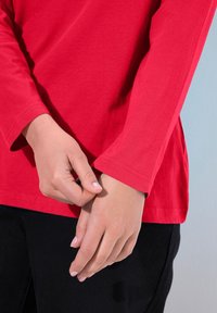 Rotes Langarm-T-Shirt mit Rundhalsausschnitt, das eine glatte Textur und einen lockeren Sitz bietet, kombiniert mit schwarzen Hosen, die Hände auf den Ärmeln ruhend.