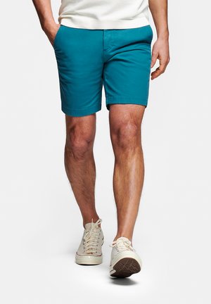 Man draagt turquoise korte broek, witte sneakers en wit shirt, loopt met één hand in de zak, staat tegen een effen witte achtergrond.