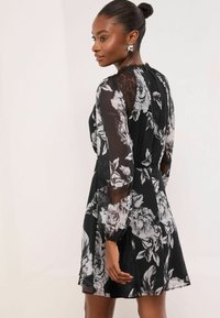 Rochie neagră cu imprimeu floral, cu mâneci lungi transparente, accente din dantelă și corsaj mulat, completată de o fustă evazată, prezentând un model cu trandafiri albi mari.