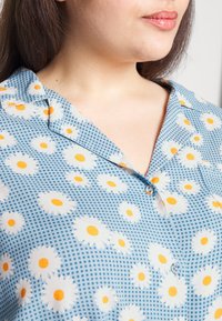 Camisa azul de botão com colarinho, apresentando um padrão xadrez e margaridas brancas com centros amarelos por todo o tecido.