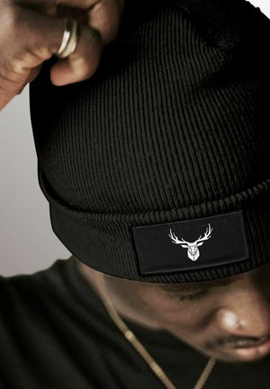 Black knitted beanie with ribbed texture. Features a rectangular black patch with a white stag logo on the front.