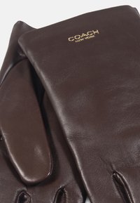 Guanti in pelle marrone con una texture liscia, caratterizzati da un design pulito e dal logo dorato "COACH NEW YORK" nell'area del polso.